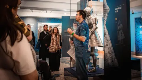 Josh Heeley/Royal Armouries A person in a blue shirt and jeans stands in front of a suit of armour in a glass case speaking to a crowd of people in a museum
