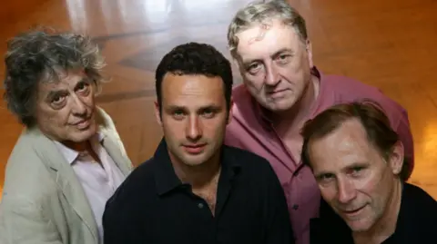 Four men stand close together and look at the camera in a head and shoulders image. Left to right, a man with grey hair, beige jacket and white shirt; a man with short dark hair and black shirt, a man with grey hair and pink shirt, and a man with brown hair and a black shirt. 