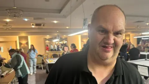 A man with very short cropped fair hair is staring into the camera. He is wearing a dark polo shirt. There are a few pool tables in the background with lots of people standing around.