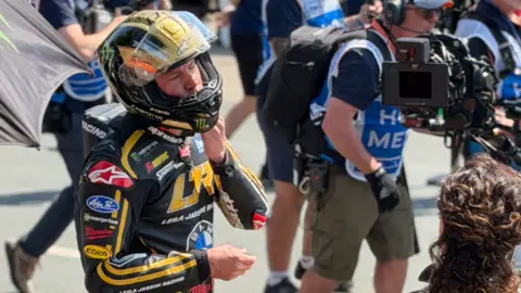 Frank Schuengel Channing Tatum is wearing black and gold leathers and a matching helmet. There are people walking behind him and a film camera facing him to the right.