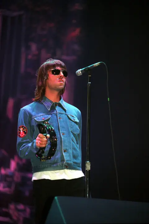 Getty Images Liam Gallagher performing at Glasgow Green, on stage wearing a blue denim jacket holding a tamborine