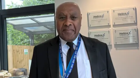 BBC A man wearing a black jacket and tie, white shirt and blue lanyard is looking at the camera. He is standing near a wall and a window, with a wooden fence behind it, is visible on the left of the photo.