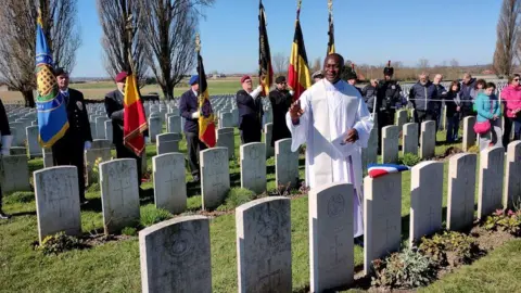 Crown Copyright A priest in white robes stands among rows of headstones in a cemetery. Behind him are military personnel holding flags and other onlookers. The sky is blue.
