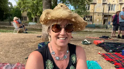 Tina Hamer wearing a straw hat and a blue necklace with a smile as she looks at the camera/ Behind her there is a tree and people on seats and standing near picnic blankets