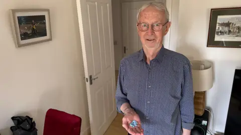 George King/BBC Tim Strugnell wearing a short and glasses and standing in his living room. He is holding out his hand on which there are several large blue tablets.