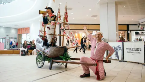 Two people dressed as pirates in a shopping centre. One is wearing a red and white, striped outfit and is kneeling on the floor, pointing to the other pirate, who is stood on a wooden ship. He is wearing white trousers, a navy and gold jacket, a black hat, and is holding a gold megaphone to his mouth.