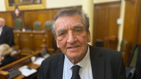 A man looking straight at the camera. He is wearing a black suit with a white shirt and black tie and is standing in front of wooden benches in a meeting room.