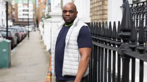 Facundo Arrizabalaga/MyLondon Darren Skeete wears a navy blue top with light green trimming round the neck and a white padded waistcoat. He is standing by black railings on a street with flats and parked cares behind him. He has a thoughtful expression.