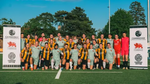 Cambridge United Women's football players from Cambridge United are wearing black and amber outfits while those from the university wear light blue. They are posing together with banners showing the logos of both establishments and the new "birthplace" logo.