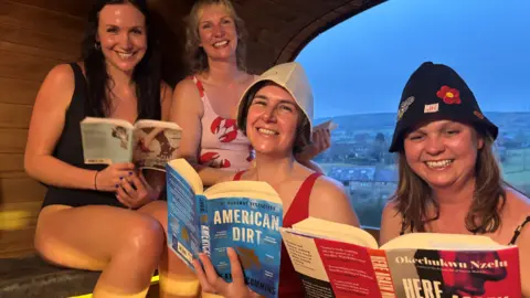 Four smiling women in their swim suits sat in a a wooden hut with a window overlooking hills and houses.