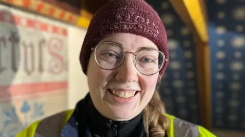 A photo of Mary who is wearing a yellow high-vis jacket, a purple hat and glasses. 