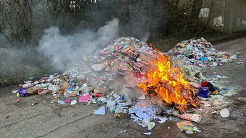 A large pile of rubbish dumped by the side of the road as a small fire burning on part of its surface. The rubbish is multi-coloured and largely appears to be discarded plastic