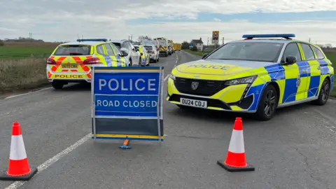 Emergency services are parked on the left side of the road and there is one police car on the right. There is a sign which says POLICE ROAD CLOSED in the middle.