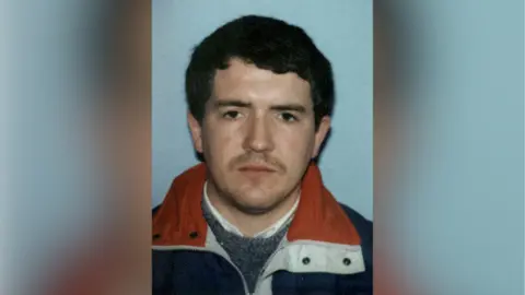 Fergal McCusker has thick brown hair parted to the side and has heavy stubble. He is wearing a blue coat, with a white and red collar and a grey jumper beneath it.