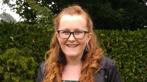 A head and shoulders shot of Amy Cooper stood in a garden, smiling at the camera. She has long, curled, ginger hair and is wearing black-rimmed glasses, silver, dangly earrings and a black leather jacket.