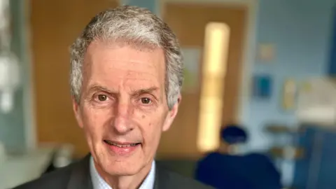 Martin Giles/BBC UEA vice‑chancellor Professor Maguire wearing a grey suit, blue check shirt and tie looking at the camera.