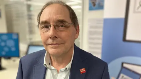 Prof Richard Luxton, Professor of bioscience and technology at UWE, smiling into the camera. He is wearing a white shirt and dark blue jacket and is sitting in a medical consultation with screens in the background