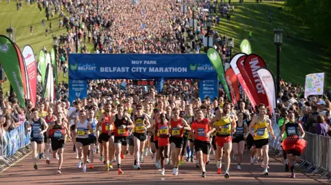 Pacemaker A large crowd of runners begin their race from the starting line at the Stormont estate. The runners, wearing athletic gear, are running underneath a large blue banner reading 'Moy Park Belfast City Marathon' and are heading towards the camera. There are supporters behind metal barricades on either side holding up supportive signs.
