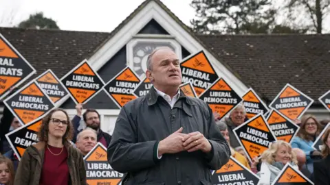 PA Media Liberal Democrat Leader Ed Davey in a wax jacket in front of a number of orange Liberal Democrat diamond-shaped placards