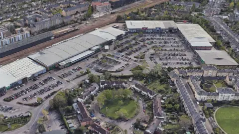 A bird's eye view of the rooftops of the some large commercial buildings next to a sizeable car park, train tracks and terraced homes, flat complexes and green spaces.