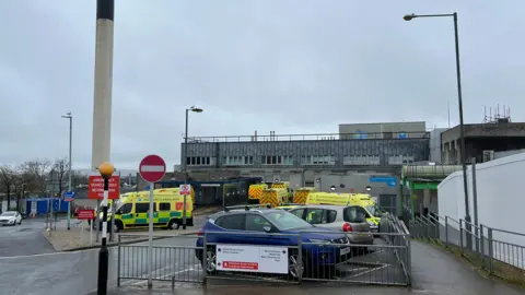 Exterior of a hospital building. The square building is large and grey. Ambulances are parked in the spaces out the front. A blue car is also parked in the car park. The sky is grey. A tall cylinder structure is built next to the main hospital building. It is white with a black top.