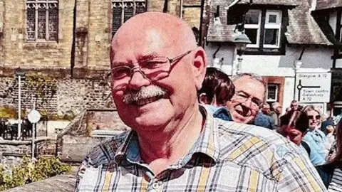 Peter Maudsley A bald headed man in a checked shirt with glasses and a moustache stands on a bridge on a sunny day