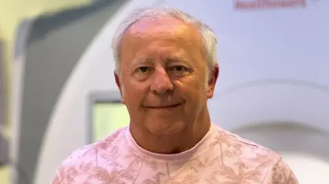 A man with short grey hair. He is wearing a pink top with palm trees on and is smiling.