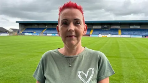 A woman with short, spikey and pink hair smiles into the camera. She is wearing a green t-shirt with a white love heart. She is standing in front of a football pitch and a seated stand with blue seats.