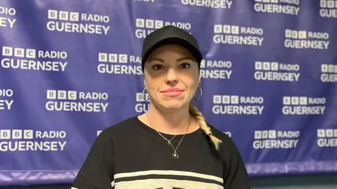 Ania Pawleta looking at the camera, with a BBC Guernsey banner behind her. She is wearing a black andgrey jumper, a necklace with a pendant, and a black cap. She has blonde hair in a ponytail draped on her left shoulder.