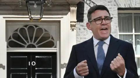 Jeff Overs/BBC Burnham speaks and gestures with his hands, with the door of Number 10 downing street just behind him. He wears a dark suit, white shirt, blue tie and square black glasses.