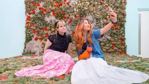two women take a selfie in front of a knitted blanket depicting features of a garden