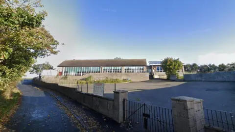 Google Slains School, exterior shot of rural school building and playground next to a country road and trees, under a blue sky.