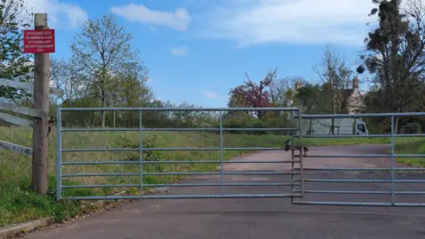 Daniel Mumby A fence across a road which blocks off access to the site. In the distance is a lorry and the house. The road is flanked by green grass.