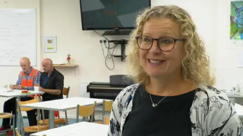 BBC Ruth has blonde hair and is wearing black glasses and a dark top