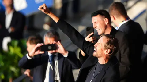 PA Peter Crouch fist bumps the air as two men take a picture of him on phones