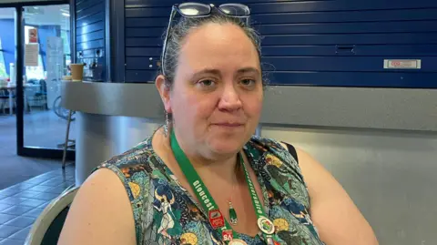 Becky Pain-Tolin sat down with a colourful dress and a green lanyard around her neck. She has glasses on her head. 