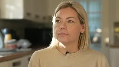 Jenny Barker sits in the kitchen wearing a cream jumper. She has shoulder-length blonde hair and a stud below her lower lip.