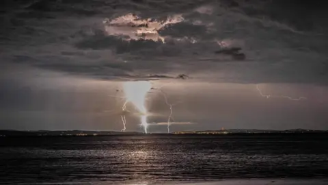 Owen Anderson Lightning pictured in the distance coming from a grey cloudy sky. The sea is at the bottom of the photo.