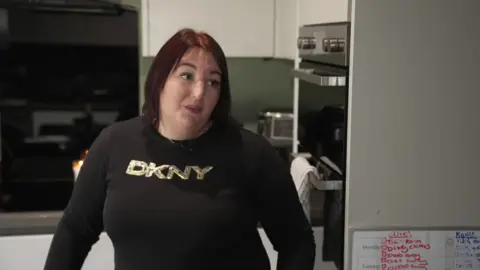 Nicha has shoulder-length dark red hair, she wears a black long-sleeved top and stands in her kitchen