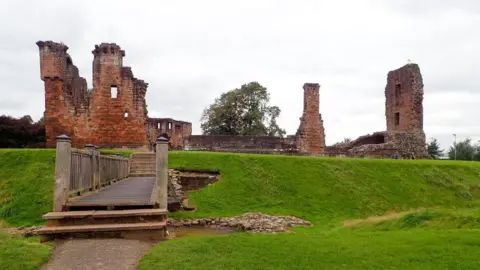 Penrith Castle. A wooden bridge leads over a grass moat to the castle ruins. The castle has no roof and only parts of two of its walls remain sanding. It is made from red brick.