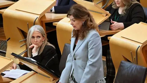 Getty Images Ruth Charteris KC and Dorothy Bain KC in the Holyrood chamber. Charteris has blonde hair and is wearing a black blazer, white blouse and glasses. She is sitting down. Bain is standing. She has brown hair and is wearing a blue jacket and floral skirt.