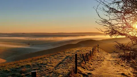 Weatherwatchers/Barnaby Bear A spectacular winter's morning with the sun's orange rays illuminating the hills near Devizes. In the foreground, a set of rickety wooden fence posts appear dark against the frosted walkway.
