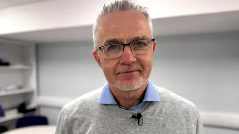 Michael McGoldrick has short grey hair and a short grey beard. He is wearing glasses, a blue shirt and a light grey jumper.