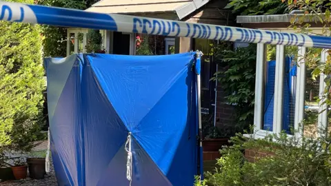 A large blue police tent, outside a house, with police tape across the front of the building. The house is brick, with lots of foliage. 