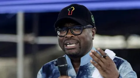 Benin s Finance Minister Romuald Wadagni in a blue shirt and a black cap, holding a microphone