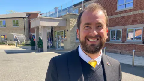 Tim wears a dark blazer with a v-neck jumper and yellow tie.
