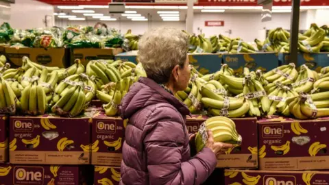 Bloomberg via Getty Images A woman holds bananas while walking through a grocery store aisle.