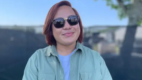 Kenneth has straight brown hair just above his shoulders and is wearing sunglasses, and a blue t-shirt under a green shirt. It is a sunny day.