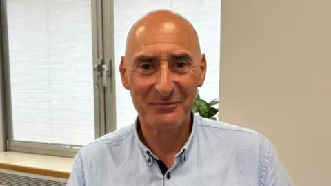 A middle aged man with a shaved head smiles into the camera. He is wearing a blue shirt, and there is an office window in the background.