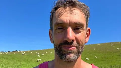 Nature Friendly Farming Network member Rhodri Lloyd-Williams stands in a field of sheep on his farm near Talybont, Ceredigion.  He has short, dark hair and a beard and is wearing a pink vest.  He is seen standing against a backdrop of a green field and blue sky.
